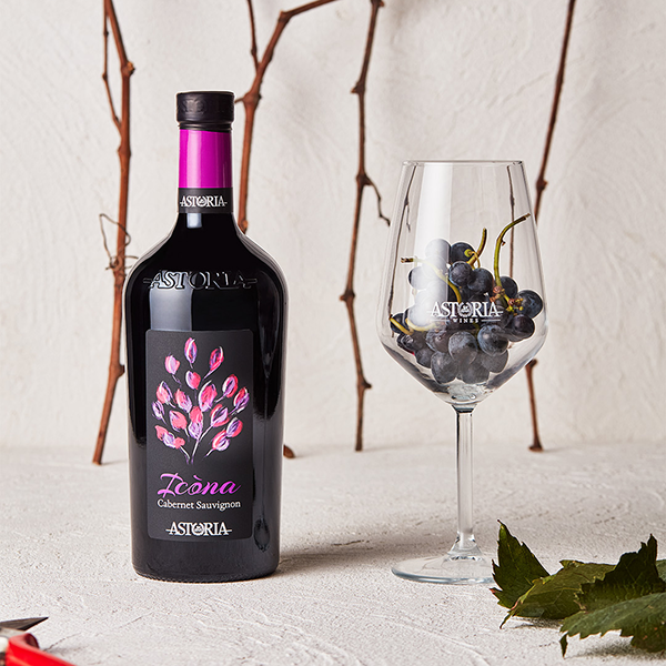 Botella de vino tinto Icona Cabernet Sauvignon de Astoria Wines junto a una copa con uvas sobre fondo natural