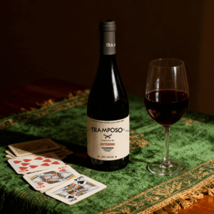 Botella de El Tramposo Sietededos Mencía junto a una copa de vino tinto y una baraja española sobre mesa verde.